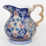 Jarra executada em porcelana, corpo globular assentado sobre base anelar, colo estreitado e borda recortada com vertedouro pronunciado, asa lateral moldada em volutas, com aplicação de douração(apresenta leves desgastes). Exterior revestido por esmalte azul cobalto, com decoração em reservas e painéis ornamentais composto por motivos florais estilizados, volutas e cartelas circulares. Os campos decorativos apresentam policromia aplicada em esmaltes relevados e contornados por filetes dourados, formando composição radiais com flores, folhas e elementos geométricos. Fundo ornamentado por arabescos brancos e pontos dourados distribuídos sobre o campo azul. Interior esmaltado em branco. Medidas: Alt.: 23 x Comp.: 25 x Larg.: 19 cm.