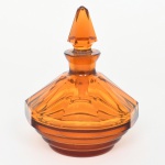 Frasco para perfume para toucador em vidro moldado e facetado, corpo de perfil octogonal, com paredes inclinadas formando facetas planas verticais que convergem para o gargalo curto e alargado. Base circular com anéis concêntricos em relevo, formando degraus horizontais. Gargalo cilíndrico com boca lapidada para encaixe de rolha maciça facetada, de formato piramidal alongado com facetas verticais e tampão cônico. Vidro translúcido em tonalidade âmbar, executada por prensagem em molde e acabamento por lapidação das facetas. Tipologia característica de vidraria decorativa do período Art Déco, com geometrização das formas e predominância de planos facetados. Apresenta quebradura na rolha, discreto bicado na ponta da tampa e marcas de uso ao redor da borda da boca. Medidas: Alt.: 14 x Diam.: 11 cm.