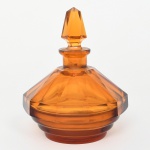 Frasco para perfume para toucador em vidro moldado e facetado, corpo de perfil octogonal, com paredes inclinadas formando facetas planas verticais que convergem para o gargalo curto e alargado. Base circular com anéis concêntricos em relevo, formando degraus horizontais. Gargalo cilíndrico com boca lapidada para encaixe de rolha maciça facetada, de formato piramidal alongado com facetas verticais e tampão cilíndrico. Vidro translúcido em tonalidade âmbar, executada por prensagem em molde e acabamento por lapidação das facetas. Tipologia característica de vidraria decorativa do período Art Déco, com geometrização das formas e predominância de planos facetados. Apresenta ínfimo trincado na rolha da tampa. Medidas: Alt.: 14 x Diam.: 11 cm.