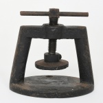 Prensa manual construída em ferro fundido, composta por base oval plana com perfuração frontal para fixação, dois montantes verticais e travessa superior. No centro da travessa encontra-se parafuso roscado vertical, acionado por manivela transversal em ferro maciço. A extremidade inferior do parafuso apresenta platô circular móvel, destinado à compressão contra a base. Estrutura monobloco em ferro fundido, com elementos mecânicos em ferro forjado e aço roscado. Medidas: Alt.: 12 x Comp.: 12 x Larg.: 9 cm.