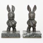 Par de esculturas confeccionadas em chumbo representando coelhos antropomorfizados, modelados em posição ereta, com orelhas longas e eretas, mãos apoiadas sobre o ventre e vestimentas estilizadas sugerindo traje infantil ou figurino de fantasia; apresentam modelagem volumétrica simples e expressão suavemente caricatural, característica de objetos decorativos do início do século XX., cada escultura encontra-se fixada sobre base retangular em mármore verde com rajados, configurando conjunto destinado ao uso como aparadores de livros (bookends). Medidas: Alt.: 15 x Comp.: 6 x Larg.: 5 cm;  Base: Alt.: 2,5 x Comp.: 10 x Larg.: 8 cm.