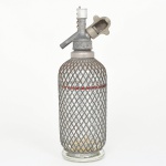 Sifão para água gaseificada, composto por recipiente cilíndrico em vidro transparente, protegido por armação metálica em malha entrelaçada, assentado sobre base circular espessa, parte superior apresenta cabeçote metálico rosqueado, com válvula de acionamento manual, bico de saída lateral e suporte para cartucho de gás, possui tampa superior com corrente metálica, elemento utilizado para retenção do componente móvel do sistema. Estrutura composta por corpo em vidro espesso soprado, grade protetora em arame metálico e mecanismo superior em metal fundido e usinado. Tipologia de sifão de soda, utilizado para carbonatação e serviço de água gaseificada ou bebidas efervescentes. Não foi testado, sem garantias de funcionamento. Apresenta marcas do tempo e de uso, visíveis nas fotografias. Medidas: Alt.: 31 x Diam.: 9 cm.