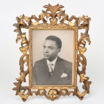 Porta-retrato em madeira ornamentada com douramento, com moldura vazada, ornamentada por motivos fitomorfos e volutas em relevo, com composição simétrica e elemento central superior em forma de palmeta. Reverso com suporte articulado em metal e placa posterior. Medidas: Alt.: x Comp.: