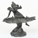 Escultura executada em metal fundido, representando figura feminina em composição alegórica, apoiada sobre elemento em forma de folhagem estilizada, integrado à base, vinculada ao estilo Art Nouveau, final do século XIX / início do século XX. Suporte usado como bandeja para correspondência, constituído por folha de grandes dimensões, com bordas recurvadas e nervuras sugeridas; base inferior com tratamento orgânico contínuo. Figura modelada em movimento, com corpo inclinado e membros projetados, trajando vestimenta com pregueado. Apresenta marcas e desgastes de uso e do tempo, demanda restauros. Medidas: Alt.: x Comp.: x Larg.: