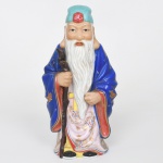 Escultura executada em porcelana representando figura de Shou Lao(shouxing), personagem da tradição chinesa associado à longevidade. Apresenta-se de pé, com barba longa e bastão sinuoso empunhado na mão direita. Traja túnica longa decoada por motivos florais estilizados e elementos fitomorfos, parcialmente recoberta por manto azul ornamentado com motivos de borboletas e pontos decorativos em reserva, cabeça coberta por gorro verde com elemento floral em relevo na parte frontal. Vestimenta inferior com padrões florais e motivos estilizados distribuídos sobre fundo claro. Base vazada na parte inferior, característica de figuras moldadas em porcelana. Medidas: Alt.: 21,5 x Comp.: 10,5 x Larg.: 7,5 cm.