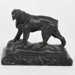 Escultura animalista executada em metal fundido, representando cão em posição de marcha, apoiado sobre base naturalista, fixada em plinto retangular em mármore. Figura modelada em volume pleno, com tratamento de superfície por cinzelamento e incisão para definição de pelagem, musculatura e feições; cabeça voltada lateralmente, orelhas pendentes, cauda curta; base superior com relevo irregular simulando terreno. Plinto em mármore de tonalidade escura com veios contrastantes, de formato retangular, com arestas retas. Medidas: