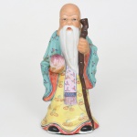 Escultura em porcelana policromada representando Shou Xing, divindade chinesa da longevidade, integrante do grupo dos Três Deuses da Fortuna (Fu, Lu e Shou). Apresenta-se de pé, longas barbas brancas e expressão serena, trajando vestes amplas ricamente decoradas com motivos florais e ornamentos lineares, segurando bastão nodoso em uma das mãos e pêssego da longevidade na outra, atributos iconográficos tradicionalmente associados a Shou Xing, as vestes incluem túnica amarela ornamentada com espirais estilizadas e manto verde decorado com pequenos ramos florais policromados, modelagem apresenta tratamento volumétrico das mangas e das dobras do traje, com barba longa moldada em relevo. A base inferior apresenta revestimento em feltro. Medidas: Alt.: 22 x Comp.: 10 x Larg.: 8,5 cm.