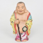 Escultura executada em porcelana moldada e esmaltada com decoração policromada representando Budai (Hotei), personagem da tradição budista chinesa associado à abundância, prosperidade e contentamento. Apresenta constituição corpulenta, cabeça raspada, expressão sorridente e ventre proeminente parcialmente exposto, trajando amplas vestes policromadas com decoração de elementos estilizados em dourado e reservas multicoloridas. Apresenta-se em posição ereta, apoiando-se em bastão nodoso, portando atributo iconográfico tradicionalmente associado a Budai; vestes apresentam manto amarelo decorado com motivos florais estilizados e estrelas policromadas, sobre túnica com reservas rosadas e faixa superior azul ornamentada com elementos dourados. A modelagem evidencia tratamento volumétrico das mangas e das dobras do traje. Apresenta perda da ponta do cajado. Medidas: Alt.: 20 x Comp.: 11 x Larg.: 8,5 cm.