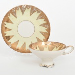 Chávena para chá em porcelana alemã, estilo Art Déco, bojo hemisférico apoiado sobre pé recortado em forma ondulada, com alça lateral estilizada, interior e aba do pires exibem decoração radial composta por reservas triangulares alternadas, preenchidas com motivos geométricos e arabescos gravados em douradura sobre fundo marfim; pires, de aba larga, repete o mesmo esquema ornamental em composição simétrica, formando padrão decorativo em raios estilizados ao redor do centro, bordas apresentam filete dourado. Marca da manufatura com indicação Bavaria, característica de manufaturas alemãs da região da Baviera. Medidas: Chávena: Alt.: 5,5 x Comp.: 13 x Larg.: 10,5 cm; Pires: Diam.: 14,5 cm.