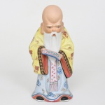 Escultura executada em porcelana representando figura de Shou Lao(Shouxing), personagem da tradição chinesa associado à longevidade, integrante do grupo conhecido como Sanxing(Fu, Lu e Shou). Apresenta-se de pé, caracterizado por crânio elevado e alongado, barba longa bifurcada e sobrancelhas arqueadas, sustenta com ambas as mão pergaminho cerimonial disposto horizontalmente à altura do torso. Traja túnica longa decorada por motivos florais estilizados e elementos fitomorfos, recoberta por manto amarelo ornamentado por motivos florais repetidos, com mangas debruadas por faixa decorativa elementos geométricos e florais. Vestimenta inferior com motivos florais e arabescos estilizados em azul e dourado, na parte posterior apresenta coque estilizado esmaltado em verde. Base vazada inferior característica de figuras moldadas. Porcelana moldada e esmaltada policromada. Manufatura chinesa, século XX. Medidas: Alt.: 22 x Comp.: 10 x Larg.: 9,5 cm.