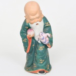 Escultura executada em porcelana moldada e esmaltada com decoração policromada representando Shou Xing (Shou Lao), divindade chinesa da longevidade e integrante da tríade auspiciosa Fu, Lu e Shou, tradicionalmente associada à fortuna, prosperidade e longevidade na iconografia chinesa. Apresenta cabeça calva de proporções acentuadas, longas barbas brancas moldadas em relevo e expressão serena, trajando amplas vestes verdes ornamentadas com motivos estilizados dourados e pequenas reservas pontilhadas. Apresenta-se ligeiramente inclinado para a frente, segurando em uma das mãos pêssego da longevidade (shoutao) e na outra tartaruga estilizada, atributos iconográficos associados à longevidade na tradição simbólica chinesa, modelagem evidencia tratamento volumétrico das mangas e das dobras do traje, com detalhes policromados em vermelho, azul e dourado nas bordas internas das vestes. Medidas: Alt.: 20,5 x Comp.: 9,5 x Larg.: 9 cm.