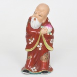 Figura em porcelana representando Shou Lao (Shouxing), divindade da tradição chinesa associada à longevidade, integrante do grupo conhecido como Sanxing (Fu, Lu e Shou). Apresenta-se de pé, com crânio proeminente e alongado, sobrancelhas arqueadas, olhos semicerrados e barba longa bifurcada em duas mechas. A cabeça apresenta leve inclinação lateral. O personagem segura pêssego da imortalidade (shoutao) junto ao peito, atributo iconográfico associado à longevidade na tradição chinesa. Traja túnica longa esmaltada em vermelho, decorada por motivos florais estilizados e representações de borboletas, executadas em esmalte policromado com aplicações em douraduras. Apresenta cabelos modelados em relevo, presos em coque posterior, e calça sapatos representados na base da túnica. Verso da base revestido com feltro. Medidas: Alt.: 20,5 x Comp.: 10 x Larg.: 9 cm.