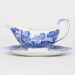 SPODE, Stoke-on-Trent - Inglaterra(fundada por Josiah Spode em 1770). Molheira executada em faiança inglesa, fixada sobre presentoir, corpo alongado de perfil naviforme, vertedouro pronunciado e alça alta em voluta. Decoração monocromática em azul sob esmalte, aplicada por técnica de transferência (transferware), representando paisagem arquitetônica de caráter pitoresco, composta por ruínas de arcadas, ponte de arco, edificações rurais, árvores e perosnagens em cena campestre, construção fortificada ou castelo ao fundo, borda do prato ondulada, ornamentada por faixa decorativa de motivos florais e arabescos estilizados, igualmente em azul. Verso apresenta marca impressa Spode, com inscrição Spode  England  Italian  Spode Design C.1816, indicando produção inglesa baseada no célebre modelo decorativo Italian, originalmente concebido no início do século XIX. Medidas: Alt.: 12 x Comp.: 22 x Larg.: 14 cm.