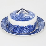 Wood & Sons, Burslem - Inglaterra(Fundada em 1865). Belíssima manteigueira em faiança inglesa da década de 1920. Esmaltagem vítrea na cor branca, dotada de profusa decoração na técnica "blue and white", intitulada 'English Scenery', com paisagem campestre contendo casario, personagem, animais, árvores e fundo com céu anuviado, tampa campaniforme apresenta alça superior arqueada, presentoir com orla circundada por flores e folhagens, borda contornada por friso na tonalidade azul cobalto. Marca da manufatura no verso.  Med.: Alt.: 8 x  Diam.: 17,5 cm. Apresenta acentuada quebradura na borda, e mancha do tempo no interior do recipiente.