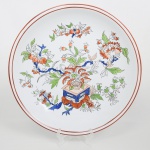 Prato medalhão executado em porcelana ao estilo Imari, decorada em policromia manual sob vidrado, apresentando composição floral de inspiração oriental, campo central ocupado por arranjo floral estilizado disposto a partir de vaso ornamental sobre suporte geométrico, do qual partem ramos sinuosos com flores abertas, botões e folhagens, executados em tons de vermelho ferroso, azul cobalto, verde e ocre claro, contornados por fino traço linear. A ornamentação apresenta clara influência do estilo Imari, tradição decorativa originária das porcelanas japonesas produzidas na região de Arita e exportadas através do porto de Imari a partir do século XVII. Esse repertório caracteriza-se pelo uso predominante da paleta azul, vermelho-ferroso e verde, bem como por composições florais e motivos vegetalistas organizados em torno de vasos e ramos estilizados. Verso apresenta anel de apoio circular elevado, típico de peças destinadas ao serviço de mesa. Medida: Diam.: 30 cm.