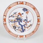 Prato medalhão em porcelana com esmaltação branca, estilo Imari de exportação, decoração em esmaltes sobre vidrado, em policromia de azul e vermelho-ferroso, com detalhes em dourado. Campo central com representação de ramo florido, com tronco sinuoso e ramificação assimétrica, apresentando flores e folhas estilizadas. Filete circular delimitando o centro. Aba ornamentada com reservas alternadas contendo motivos florais, intercaladas por padrão reticulado contínuo. Base com anel de apoio rebaixado. Medida: Diam.: