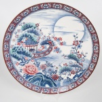 Grande prato medalhão em porcelana japonesa, estilo Imari, período Shwa, meados do século XX. Decoração em esmaltes sob vidrado, policromia em azul, vermelho e verde, com reservas em branco. Cena central com paisagem de inspiração oriental, composta por curso dágua, vegetação estilizada, flores e estrutura arquitetônica leve com cobertura reticulada. Presença de lua cheia ao fundo. Ornamentação com elementos florais e folhagens distribuídas no campo. Aba com friso contínuo em padrão repetitivo geométrico e fitomorfo em vermelho e azul. Base com anel de apoio rebaixado. Marca em vermelho sob esmalte, com caracteres orientais e inscrição Japan. Medida: Diam.: