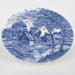 Johnson Brothers, Staffordshire - Inglaterra(Fundada em 1883). Belíssima travessa oval executada em faiança inglesa, padrão Watermill. Decoração monocromática em azul sob esmalte, representando paisagem rural com moinho hidráulico, executada por estampagem. A composição mostra edifício de moinho com roda dágua em funcionamento, pequeno dique e queda dágua, acompanhados por casas campestres, árvores frondosas, figuras humanas em ambiente bucólico e céu anuviado ao fundo; orla modelada com delicado movimento ondulado, borda recortada com arcos em repetição. Marca da manufatura no verso. Medida: Comp.: 30 x Larg.: 23,5 cm.