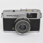Câmera fotográfica Olympus Trip 35. Câmera fotográfica compacta de filme 35 mm, com corpo metálico de acabamento prateado e revestimento frontal em material sintético texturizado de cor preta. Apresenta visor óptico frontal integrado, janela do fotômetro de selênio e conjunto de lente fixa central. Equipada com lente Olympus D.Zuiko 40 mm f/2.8, fabricada no Japão, com anel frontal graduado para controle de distância focal e anel de abertura com valores f/2.8 a f/22, incluindo posição automática para exposição. Sistema de obturador mecânico com seleção automática de exposição mediante célula fotossensível de selênio, dispensando uso de bateria. Parte superior com disparador, sapata para flash e contador de exposições; base com rosca metálica para fixação em tripé e gravação Made in Japan. Tampa traseira articulada para inserção do filme fotográfico. Não foi testada, sem garantias de funcionamento, estado conforme fotografias. Medidas: Alt.: 7 x Comp.: 12 x Larg.: 6 cm.