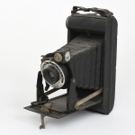 Câmera fotográfica portátil do tipo câmera de fole (folding camera), modelo Kodak Folding Brownie Six-20, fabricada pela Eastman Kodak Company, Inglaterra, c. 1934. Estrutura metálica revestida em material sintético preto, dotada de sistema de abertura frontal com fole retrátil, no qual o conjunto óptico se projeta por meio de plataforma metálica articulada quando a tampa frontal é aberta. Equipada com obturador Kodak Brownie Shutter, com posições I (instantâneo) e B (bulb), e lente menisco Kodak, com indicação de ajustes simples de foco (Kodette F11). O visor óptico reflexo lateral permite enquadramento tanto na posição horizontal quanto vertical; compartimento interno destina-se ao uso de filme em rolo Kodak 620, conforme indicação presente no interior da tampa. Parte traseira removível para inserção do filme, dotada de janela rubi para leitura da numeração das exposições. Produzida pela Eastman Kodak Company, na unidade de fabricação britânica, sendo este modelo introduzido em 1934, período em que as câmeras portáteis de fole se popularizaram entre fotógrafos amadores. Estado de conservação conforme fotografias, apresenta desgaste compatível com o tempo e uso, com marcas de oxidação superficial nas partes metálicas, desgaste no revestimento externo e sinais de uso no conjunto mecânico e rasgos no fole, mantendo entretanto a integridade estrutural do aparelho. Medida aberta; Alt.: 7 x Comp.: 13,5 x Larg.; 7,5 cm.