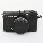 Câmera fotográfica portátil do tipo compacta para filme 35 mm, modelo Olympus Trip 35, fabricada pela Olympus Optical Co., Ltd., Japão, c. 1967. Câmera fotográfica de corpo metálico com acabamento pintado em preto e revestimento frontal em material sintético texturizado, dotada de visor óptico frontal e célula fotossensível de selênio integrada para medição automática de exposição. A parte superior apresenta disparador, sapata para flash e seletor de rebobinamento do filme. Equipada com lente Olympus D.Zuiko 40 mm f/2.8, fabricada no Japão, com anel frontal de foco graduado em metros e pés e anel de abertura com valores f/2.8 a f/22, incluindo posição automática para controle de exposição. O sistema de obturador é mecânico, associado ao fotômetro de selênio, dispensando o uso de bateria. Parte inferior com inscrição Made in Japan e rosca metálica para fixação em tripé. Compartimento traseiro articulado para inserção de filme fotográfico formato 135 (35 mm). Estado de conservação conforme fotografias, apresenta desgaste de uso e oxidação superficial nas bordas metálicas, perda parcial do acabamento externo e marcas de manuseio, mantendo contudo a integridade estrutural do corpo, da lente e do compartimento interno do filme. Não foi testado, sem garantias de funcionamento Medidas: Alt.: 7 x Comp.: 12 x Larg.: 6 cm.