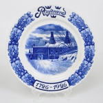 Rörstrand, Estocolmo - Suécia(fundada em 1726 pelo dinamarquês Johann Wolff). Raro prato de coleção comemorativo de 200 anos de fundação da manufatura, executado em faiança, esmaltação na cor branca, decorado em azul cobalto aplicado sob o vidrado, técnica tradicional associada às manufaturas europeias inspiradas no repertório do "blue and white"; aba ornamentada por guirlanda contínua de folhas de carvalho estilizadas, dispostas em composição simétrica envolvendo o campo central. Na parte superior encontra-se a inscrição Rörstrand, acompanhada por três coroas, elemento heráldico característico da iconografia sueca. Na parte inferior figura cartela com a inscrição 17261926, indicando a produção comemorativa do bicentenário da fundação da manufatura Rörstrand, estabelecida em 1726 e uma das mais importantes fábricas de porcelana e faiança da Escandinávia; medalhão central apresenta representação da antiga fábrica da Rörstrand, com edifícios industriais, chaminés e paisagem invernal, executada em gradações tonais de azul que conferem efeito pictórico à composição. Verso apresenta marca impressa em azul com a inscrição Rörstrand, correspondente à manufatura sueca. Medida: Diam.: 25 cm.
