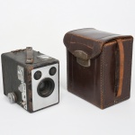 Câmera fotográfica tipo box Six-20 Brownie Model E, fabricada pela Kodak Limited, Londres, Inglaterra, c. década de 1930, acompanhada de estojo original em couro e material impresso de instruções. Câmera fotográfica portátil de corpo retangular, construída em estrutura metálica revestida externamente por material sintético texturizado; face frontal apresenta placa metálica com lente central fixa, acompanhada por duas pequenas janelas ópticas destinadas ao sistema de visor, parte superior com visor adicional e alça de transporte em couro fixada por rebites metálicos, conjunto ótico composto por lente simples de foco fixo, característica das câmeras populares da série Brownie, associada a obturador mecânico de acionamento manual, lateral dotado de botão de avanço do filme, parte posterior possui janela circular com filtro vermelho, destinada à visualização da numeração do filme durante o transporte do rolo; interior da câmera abriga compartimento para filme fotográfico formato 620, sistema amplamente utilizado pela Kodak nas câmeras box da primeira metade do século XX. Acompanha estojo original em couro rígido com fecho metálico, folhetos e material de instruções da Kodak, incluindo manual intitulado How to use your Six-20 Brownie Model E. Estado de conservação conforme fotografias, presenta marcas de uso, desgaste do revestimento externo, oxidação superficial em partes metálicas e envelhecimento natural do estojo de couro, mantendo contudo sua estrutura completa, com componentes principais preservados e acompanhada de material impresso original. Não foi testada, sem garantias de funcionamento. Medidas: Alt.: 10 x Comp.: 12 x Larg.: 8 cm.