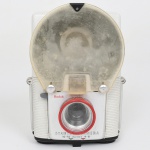 Câmera fotográfica infantil Kodak Startech Camera com flash integrado, fabricada pela Eastman Kodak Company, Estados Unidos, c. década de 1950. Câmera fotográfica do tipo box camera destinada ao público infantil, confeccionada em corpo de material plástico moldado na cor branca com base inferior em material sintético preto. O modelo apresenta flash incorporado em refletor metálico superior, protegido por difusor plástico translúcido removível, destinado à dispersão uniforme da luz durante a exposição. A parte frontal exibe inscrição Kodak e denominação Startech Camera, com indicação de distâncias de foco 48 inches e 1016 inches, correspondentes a fotografias de curta distância, característica comum em câmeras destinadas a uso recreativo infantil. O conjunto ótico é composto por lente simples fixa com aro frontal em plástico vermelho, associada a visor óptico superior. O compartimento posterior abriga mecanismo para filme fotográfico em rolo, com carretel removível e sistema manual de avanço do filme. Na parte traseira encontra-se etiqueta do distribuidor Lester A. Dine Inc., New Hyde Park, Long Island, New York, empresa conhecida pela distribuição de equipamentos fotográficos e didáticos nos Estados Unidos. Estado conforme fotografias, com marcas de uso, pequena avarias e perdas. Não foi testada, sem garantias de funcionamento. Medidas: Alt.: 13,5 x Comp.: 9 x Larg.: 6 cm.