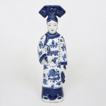 Escultura em porcelana chinesa azul e branca representando dama cortesã. Figura feminina em posição ereta, trajando vestes cerimoniais ricamente decoradas com motivos de dragões imperiais e nuvens auspiciosas, elementos tradicionais da iconografia chinesa associados a poder e boa fortuna. A personagem apresenta expressão serena, cabelos presos em elaborado coque encimado por amplo toucado estilizado, e segura em uma das mãos um cachimbo longo, acessório característico de distinção social na cultura chinesa tradicional, superfície decorada em esmalte azul cobalto sobre fundo branco, evocando a tradição da porcelana clássica produzida em Jingdezhen, célebre centro cerâmico da China, base apresenta selo quadrangular em caracteres chineses em relevo, em estilo arcaizante inspirado em marcas imperiais da dinastia Qing. Medidas: Alt.: 34 x Comp.: 10,5 x Larg.: 7,5 cm.