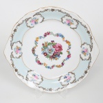 VIZAVI. Grande prato para serviço em porcelana, esmaltação branca parcialmente preenchida por faixa de tonalidade azul claro, dotado de rica decoração com flores e elementos vegetalistas em estampagem policromada, bordo recortado contornado com filete em douradura, relevado com volutas e conchas, alças laterais integradas. Marca da manufatura no verso(sobreposição). Medidas: Comp.: 28 x Larg.: 26 cm.