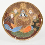 Prato circular em cerâmica japonesa com decoração policroma, aplicação de moriage em relevo, realces em ouro e filete na borda, dotada de pequenas alças integradas; face frontal apresenta composição figurativa formada por uma figura central sentada, acompanhada por quatro Imortais (sennin/rakan), representados como anciãos em trajes tradicionais, tema recorrente em produções de exportação em estilo Satsuma. O fundo do campo decorativo é preenchido por padrão de ondas, com áreas delimitadas por contornos dourados. A iconografia e a técnica de moriage situam a peça dentro da produção japonesa voltada ao mercado externo entre o final do período Taish e o início do período Showa. Apresenta craquelado característico do material acentuado pelo tempo, e dois fios no verso da borda. Medida: Diam.: 24,5 cm.