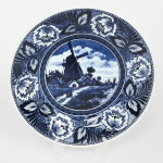Boch Frères Keramis (posteriormente Royal Boch), La Louvière - Bélgica(Fundada em 1841 por Jean-François Boch). Prato de coleção em faiança esmaltada, de formato circular com aba larga e cavidade rasa, decorado na técnica de transferência (transferware) em azul sobre fundo branco; cena central apresenta paisagem rural de inspiração holandesa, com moinho de vento, casa, curso dágua e fundo com céu anuviado, em composição monocromática pontilhada, orla circundada por faixa contínua com motivos florais estilizados e elementos vegetalistas, organizados em repetição, delimitados por frisos concêntricos. Verso com marca impressa contendo inscrição BOCH Belgium, acompanhada de elemento heráldico. Medida: Diam.: 23,5 cm.