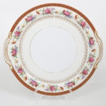 NORITAKE, Nagoya - Japão(Fundada em 1904 pelos irmãos Ichizaemon e Toyo Morimura). Exuberante travessa circular executada em porcelana japonesa, esmaltagem vítrea na cor branca, orla circundadas por buquês de flores em estampagem policromada sobre fundo levemente em tom marfim, dotada de profusa ornamentação em douradura, com frisos, arabescos, e elementos vegetalistas sobrepondo faixa de tonalidade bordô contornando a borda, abas laterais proeminentes, recortadas e terminadas em volutas. Marca da manufatura ao fundo. Med.: Diam. 27,5 cm. Apresenta desgastes parciais na douradura.