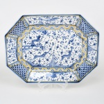 Travessa em faiança, produção portuguesa da região de Coimbra, estilo barroco de influência seiscentista. Forma oitavada com bordo recortado e aba facetada. Decoração em azul sobre fundo branco, com aplicação de filetes em amarelo. Campo central com composição de animais estilizados, incluindo quadrúpedes em movimento, aves e elementos fitomorfos distribuídos em padrão contínuo. Presença de elemento arquitetônico estilizado ao centro. Aba segmentada com reservas decoradas, alternando painéis com motivos vegetalistas e padrões geométricos, incluindo escamas e volutas.  Verso contendo inscrição manuscrita em azul sob esmalte: Atelier B.L., Coimbra, Portugal, Séc. XVII, indicativa de reprodução historicista. Medidas: Comp.: 33 x Larg.: 25 cm.