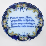 Prato medalhão de parede em faiança, produção portuguesa da região de Alcobaça; formato circular de borda modelada com ondulações, contornos em tonalidade azul cobalto e centro em azul claro, orla envolta por volutas e padrão de flores em policromia, centralizando verso com mensagem auspiciosa: "Plena de graça, Maria, Virgem-Mãe do Redentor, Enchei sempre de alegria Nosso Lar, ninho de amor". Verso com iniciais do fabricante, e localizado Alcobaça-Portugal. Apresenta perfurações no verso, para encaixe de suporte com a finalidade de afixação em parede. Borda com dois desgastes na esmaltação. Medida: Diam.: 30 cm.