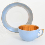 Porcellana Mauá, Mauá - SP(Fundada em 1937 por Fritz Erwin Schmidt / ativa até 1968). Xícara de coleção, para café, em porcelana nacional da década de 1940/50; esmaltagem de tonalidade azul claro, bojo hemisférico de interior integralmente preenchido com douradura, alça lateral em voluta e borda do pires ondulada, ambas contornadas por filete dourado. Marca da manufatura no verso. Medidas: Xícara: Alt.: 4 x Comp.: 8,5 x Larg.: 6,5 cm; Pires: Diam.: 10,5 cm. Apresenta leves desgastes parciais na douradura causada pelo uso.