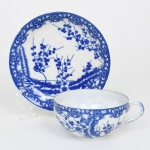 Xícara para chá em porcelana japonesa, dita "casca de ovo", esmaltação branca, estilo Arita de inspiração blue and white, período Shwa, meados do século XX; bojo hemisférico, alça lateral em voluta; decoração em azul sob vidrado, com composição de ramos floridos estilizados, distribuídos em paisagem contínua. Bordas com friso ornamental em padrão repetitivo de volutas e elementos fitomórficos. Marca da manufatura no verso. Medidas: Xícara: Alt.: 4,5 x Comp.: 10,5 x Larg.: 9 cm; Pires: Diam.: 13 cm.