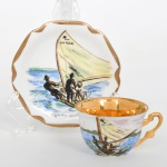 Xícara de coleção, para café, em porcelana nacional, esmaltação branca, interior do bojo integralmente preenchido em douradura, decoração em pintura manual sobre vidrado, com composição narrativa de cena marítima: jangada com dois pescadores em ação, conduzindo jangada pelo vento, sobre faixa dágua em tons de azul, com linha de horizonte sugerida ao fundo. Inscrições Do Mar na vela e Jangada Bahia na reserva do pires. Paleta com variações de azul, verde, ocre e vermelho. Medidas: