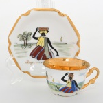 Porcellana Mauá, Mauá - SP(Fundada em 1937 por Fritz Erwin Schmidt / ativa até 1968). Xícara de coleção, para de café, em porcelana nacional da década de 1950/60, esmaltação branca, interior do bojo integralmente preenchido em douradura, decoração em pintura manual sobre esmalte, com composição figurativa de cena regional: figura feminina em traje típico, com torso inclinado e braço elevado sustentando recipiente sobre a cabeça, disposta em paisagem litorânea com faixa de água, vegetação e embarcação ao fundo. Inscrição Negrinha  Bahia na reserva do pires.  Verso com marca da manufatura em verde sob vidrado e inscrição manuscrita Pintado à mão. Medidas: