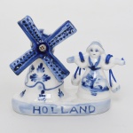Miniatura decorativa em porcelana holandesa Delft, representando moinho e figura feminina tradicional, esmaltação branca, modelado sobre base oblonga com inscrição frontal HOLLAND,  apresenta moinho de vento estilizado com pás móveis, decorado em azul cobalto sob o vidrado, acompanhado por figura feminina trajando vestimenta tradicional holandesa.A personagem é representada com vestido típico e chapéu característico, apresentando braços  com cordões têxteis azuis, elemento comum em pequenas lembranças turísticas produzidas na Holanda. O moinho apresenta decoração floral estilizada e detalhes pontilhados em azul, evocando a tradição da porcelana e faiança Delft. Verso traz marca em azul Handpainted in Delft blue colour  E H  10412, indicando produção decorativa inspirada na estética Delft, pintada manualmente. Medidas: Alt.: 8,5 x Comp.: 9 x Larg.: 4,5 cm.