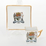 Porcelana Condessa S/A, Blumenau - SC.(Fundada por Nicolas Coman, e Hercílio Deeke, em 1956/ ativa até 1967). Xícara para café em porcelana nacional da década de 1950/60; esmaltação na cor branca, bojo quadrangular com boca ligeiramente ondulada e asa lateral estilizada; pires de formato quadrangular com aba recurvada. Decoração central aplicada por decalque com brasão do município de Blumenau, encimado por coroa mural e ladeado por duas figuras alegóricas: à esquerda Dr. Hermann Bruno Otto Blumenau (18191899), fundador da colônia de Blumenau, representado em traje civil do século XIX; à direita figura masculina representando colono agrícola, trajando vestimenta de trabalho e portando instrumento agrícola. Sob o escudo, listel com a inscrição PRO SANCTA CATHARINA ET BRASILIA e identificação BLUMENAU. Bordas da xícara e do pires com filete dourado. Marca da manufatura no verso. Medidas: Xícara: Alt.: 5 x Comp.: 6 x Larg.: 4,5 cm. Pires: Comp.: 9 x Larg.: 9 cm.