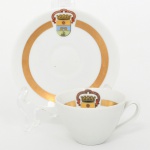 Porcelana Renner, Porto Alegre - RS(Fundada em 1937). Elegante xícara de coleção, para café, em porcelana nacional. Padrão Donaire; esmaltação na cor branca, decorada com faixa circular em douradura junto à borda da xícara e no centro do pires. Apresenta brasão heráldico da Cidade de Porto Alegre, encimado por coral mural e ladeado por listel contendo a inscrição "Leal e Valerosa Cidade de Porto Alegre", aplicado em estampagem policromada. Escudo com campo tripartido: faixa superior amarela com Cruz da Ordem de Cristo, campo central azul com portal de cantaria, campo inferior verde com embarcação à vela sobre águas. Marca da manufatura no verso. Medidas: Xícara: Alt.: 4,5 x Comp.: 8 x Larg.: 6,5 cm; Pires: Diam.: 10,5 cm.