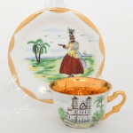 Xícara de coleção, para café, em porcelana nacional, esmaltação branca, interior do bojo integralmente preenchido em douradura, decoração policroma em pintura manual sobre esmalte, com composição figurativa e arquitetônica. Na xícara, representação da Igreja do Senhor do Bonfim, com fachada, torres sineiras laterais e frontão central, inserida em paisagem com vegetação estilizada. No pires, cena regional com figura feminina trajando indumentária típica, disposta em paisagem litorânea com palmeira, linha dágua e embarcação ao fundo. Inscrição Bahia na reserva do pires. Verso com inscrição manuscrita Pintado à mão. Medidas: