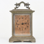 Ansonia Clock Company, Connecticut, EUA(Fundada em 1851 por Anson Green Phelps).Relógio de viagem/despertador mecânico, fabricado pela histórica fabricante norte-americana Ansonia Clock Company, em caixa metálica fundida (provavelmente liga de zinco ou spelter) com acabamento prateado envelhecido e decoração em relevo de inspiração Art Nouveau / Edwardiana, composta por frisos vegetalistas e arabescos. Caixa apresenta formato retangular com cantos suavemente arredondados, base moldurada ornamentada e alça superior vazada com volutas e elementos estilizados, apoiada sobre dois pinos esféricos. Mostrador de papel sobre chapa metálica, com numerais arábicos em preto, ponteiros estilizados em aço e pequena sub-esfera de segundos posicionada às seis horas; conjunto protegido por vidro frontal bisotado encaixado em moldura metálica. No campo central do mostrador aparece a inscrição da marca Ansonia. Movimento interno mecânico, construído em latão, composto por platinas recortadas com visores para observação da engrenagem, regulador superior marcado FS (Fast / Slow) para ajuste de marcha e conjunto de molas reais alojadas em tambor, responsável pelo funcionamento do relógio e pelo sistema de soneria. Fundo da caixa possui tampa metálica que permite acesso ao mecanismo para manutenção, lubrificação e ajuste; produção industrial norte-americana do início do século XX (aprox. 19001920), período em que a Ansonia produziu grande quantidade de relógios domésticos decorativos para exportação. Exemplar em estado de conservação compatível com a idade, apresentando oxidação superficial, desgaste da pátina e acúmulo de poeira no movimento, porém preservando mostrador original, ponteiros e estrutura completa. Não está funcionando, não foi testado, sem garantias de funcionamento. Medidas: Alt.: 19,5 x Comp.: 12,5 x Larg.: 8,5 cm.