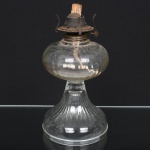 Candeeiro de mesa a querosene, executado em vidro prensado, translúcido e incolor, composto de reservatório globular, de formato abaulado, apresentando pequenas saliências circulares em relevo distribuídas pelo bojo, características do processo de moldagem industrial do vidro, reservatório apoiado sobre coluna curta em vidro liso que se desenvolve para base campaniforme alargada, moldada com caneluras verticais radiais, formando pé largo destinado a conferir estabilidade ao conjunto; parte superior dotado de queimador metálico para pavio, em liga metálica atualmente com pátina de oxidação compatível com a idade, dotado de roda lateral dentada para regulagem do pavio e galeria com suportes curvos destinados à fixação da chaminé de vidro (ausente). O sistema permitia a elevação e ajuste do pavio, controlando a intensidade da chama alimentada por querosene armazenado no reservatório. Peça de produção utilitária seriada, típica do período anterior à difusão generalizada da iluminação elétrica, fabricada entre o final do século XIX e as primeiras décadas do século XX, exemplar preserva o reservatório, base e queimador originais, apresentando sinais de uso e envelhecimento natural, pequenas bolhas e irregularidades no vidro decorrentes do processo de prensagem e ausência da chaminé superior. Provável datação c. 19001930. Medidas: Alt.: 27,5 x Diam.: 14 cm.