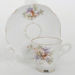 Chávena de coleção, para café, em porcelana europeia do início do século XX, período e estilo Art Nouveau; esmaltação na cor branca, modelada com elementos do gênero sob o vidrado, decorada com composição botânica em policromia, predominando as tonalidades violeta, verde e marsala, realçado com singelos arremates em douradura(apresenta desgaste); bojo obcônico facetado com oito lados, base com pequenos apoios moldados e asa lateral estilizada, padrão decorativo replicado no pires. Marcado com numeração ao fundo. Medidas: Alt.: 5,5 x Comp.: 7,5 x Larg.: 5,5 cm; Pires: Diam.: 11 cm.