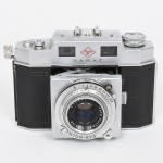 Câmera fotográfica de médio formato Agfa Karat. Corpo em metal com revestimento em material sintético preto texturizado, de construção retangular com arestas facetadas. Parte superior em metal com inscrição Agfa Karat e duas janelas do visor frontal. Equipada com objetiva Agfa Solinar 1:2.8 f=50 mm, montada em conjunto Prontor-SVS, com anéis graduados para controle de velocidade do obturador e abertura do diafragma. Sistema de foco frontal com escala graduada, montado em suporte retrátil característico das câmeras da série. Interior com compartimento para filme em cartucho, apresentando numeração RN 1564. Base com inscrição Made in Germany. Acompanha estojo original em couro marrom, com costuras aparentes e gravação Karat 6015  Made in Germany. Estado conforme fotografias, não foi testada, sem garantias de funcionamento. Medidas: Alt.: 9,5 x Comp.: 13,5 x Larg.: 7,5 cm.