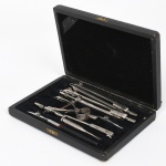 Estojo contendo conjunto de instrumentos para desenho técnico, acondicionados em caixa retangular em madeira revestida, interior forrado em veludo escuro, com tampa articulada e fecho metálico. Compartimento único com encaixes para acondicionamento dos instrumentos.Conjunto composto por compassos de diversas dimensões, tira-linhas, extensores, pontas intercambiáveis e adaptadores. Peças em metal, com hastes de seção cilíndrica e superfícies estriadas para empunhadura; parafusos de ajuste micrométrico e articulações rebitadas. Presença de suporte circular para fixação de grafite ou pena. Apresentam sinais de oxidação nos metais, desgaste do revestimento interno. Medidas: