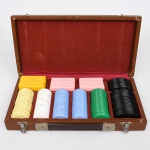 Conjunto de fichas para jogo, confeccionado em material sintético, acondicionado em estojo retangular em madeira, com tampa articulada, fechos metálicos frontais e interior compartimentado revestido em tecido vermelho; compartimentos internos destinados à organização das fichas por cor e valor. Conjunto composto por fichas circulares e retangulares em diferentes cores (branco, amarelo, azul, verde, preto e rosa), com valores em relevo, incluindo 20, 50, 200, 500 e 1000.