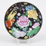 Tigela executada em porcelana com montagem metálica, apresentando decoração policromada sobre fundo preto, composta por exuberante arranjo de flores, frutos e elementos vegetais estilizados em policromia; contendo peônias, rosas, lótus, ramos floridos e cachos de uvas, acompanhados por frutos como romã e cítrico, executados em esmaltes nas tonalidades de rosa, azul, amarelo, verde, branco e laranja, com contornos definidos por traço escuro, composição decorativa ocupa integralmente o campo interno do prato, formando arranjo assimétrico de caráter naturalista, típico de produções decorativas chinesas destinadas à exportação durante o século XX. O fundo de tonalidade preta intensifica o contraste cromático dos esmaltes policromos, característica frequentemente empregada em porcelanas decorativas desse período. Verso apresenta estrutura metálica de suporte e suspensão, permitindo a fixação do prato em parede para uso ornamental. No centro do fundo encontra-se marca circular em esmalte vermelho, com a inscrição: Decorative by Noritake  Sent by S. I. & Co.  Made in China (algumas letras apresentam desgaste natural).A marca indica produção chinesa para exportação, provavelmente vinculada a distribuição ou decoração comercializada por empresa associada à marca Noritake, prática comum em peças decorativas do século XX. Medida: Diam.: 18 cm.